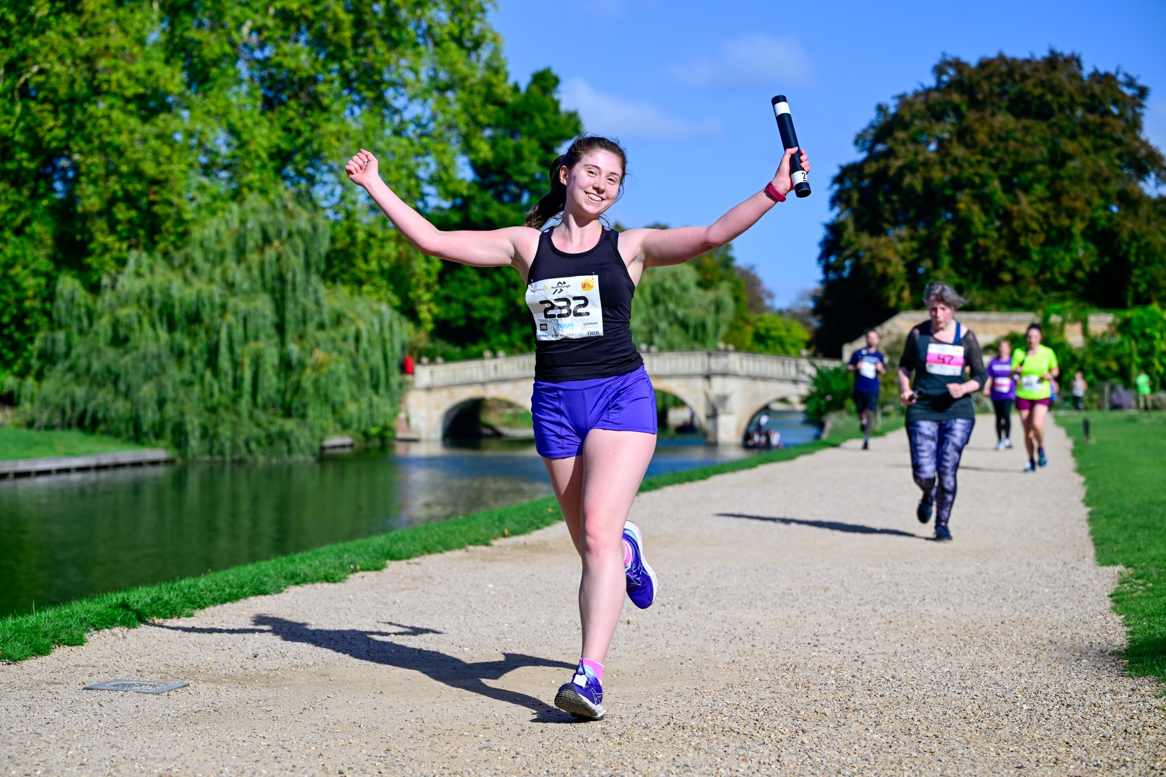 Runner carrying a relay baton along a riverside route in Cambridge during Chariots of Fire, with fellow participants running behind.